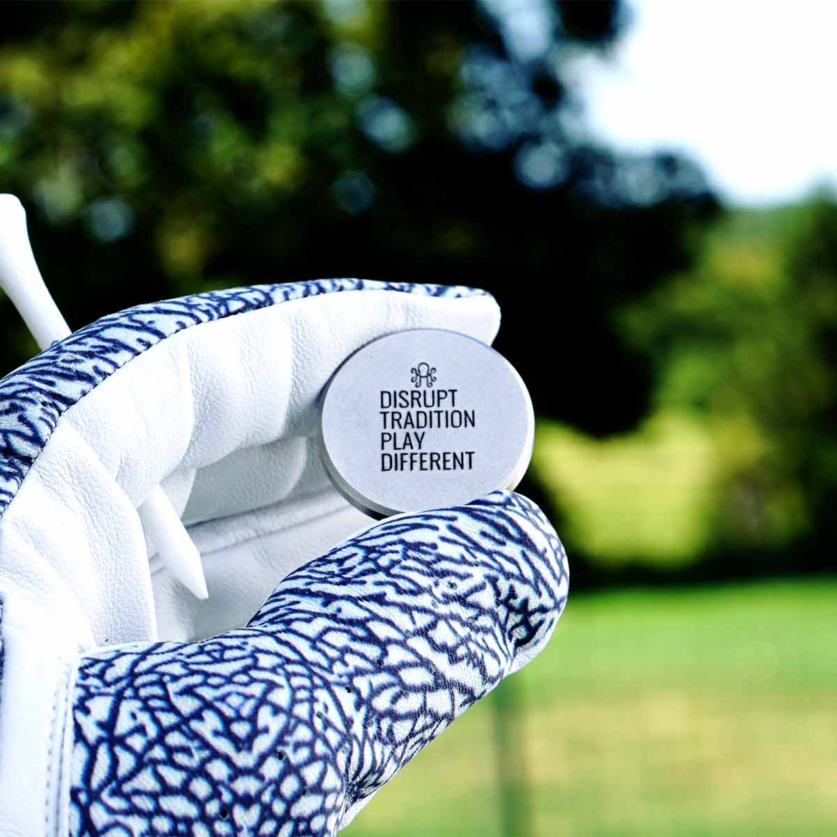 Golf glove holding a golf ball marker with a 'Disrupt Tradition, Play Different' message against a blurred green background.