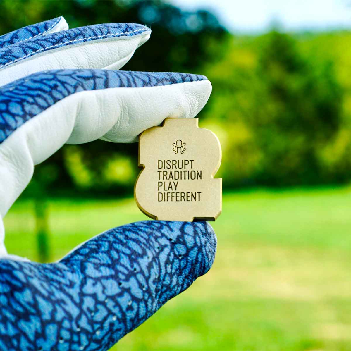 Golfer wearing a golf glove holding a brass money sign shaped golf ball marker, outdoors.
