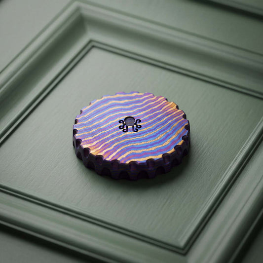 Hobo Luck Timascus golf ball marker back view with Kraken octopus cutout and scalloped edges on heat-colored layered titanium, resting on green paneling