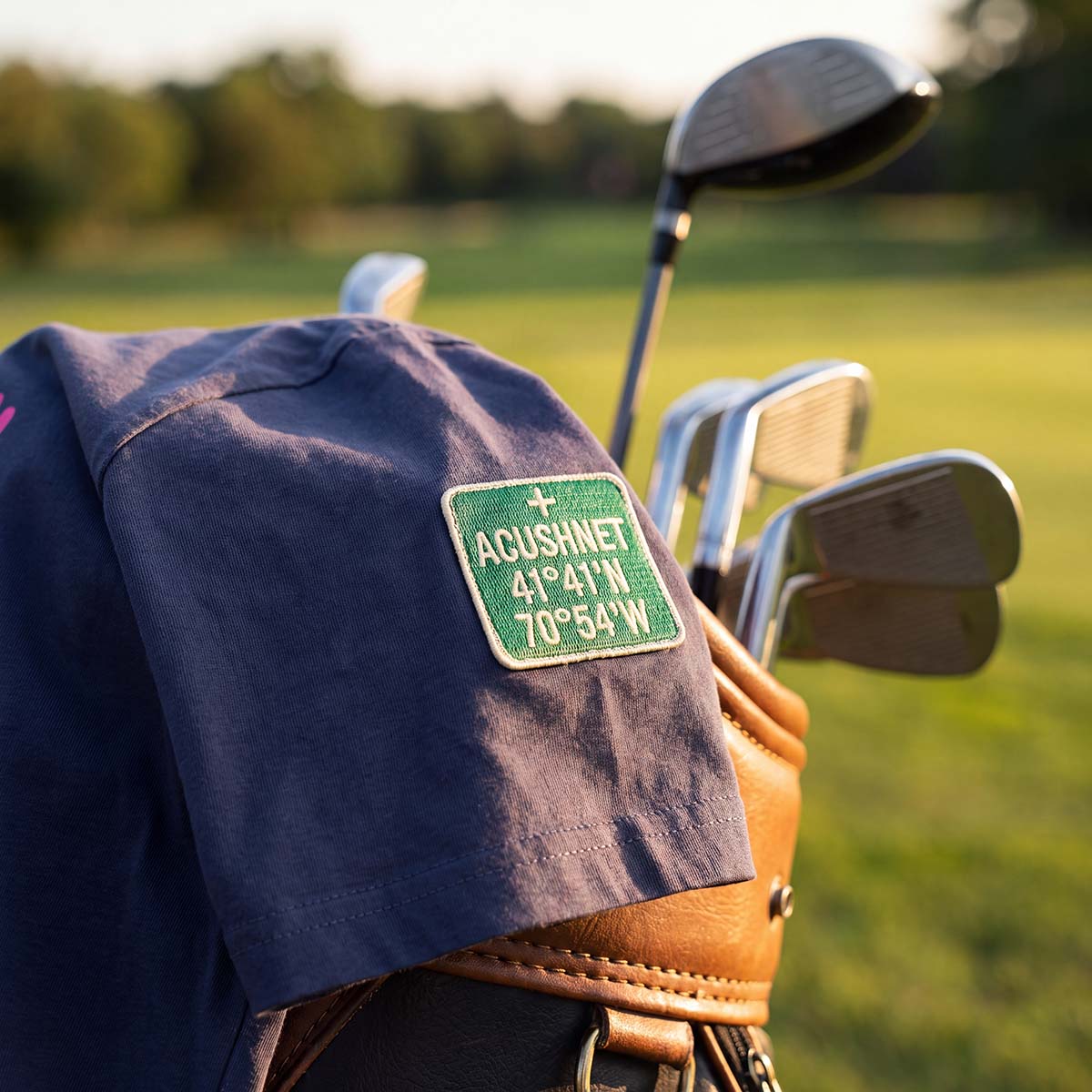 A navy blue golf shirt with a green and white 'ACUSHNET' patch on the sleeve, resting on a tan golf bag with golf clubs on a blurred green golf course background.