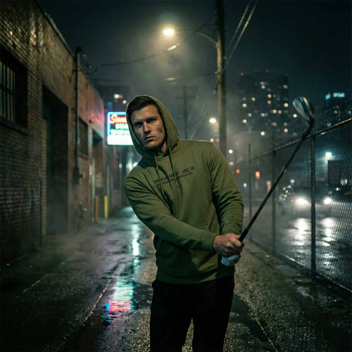 Golfer in the Garage hoodie holding a golf club on a rain-soaked street at night.