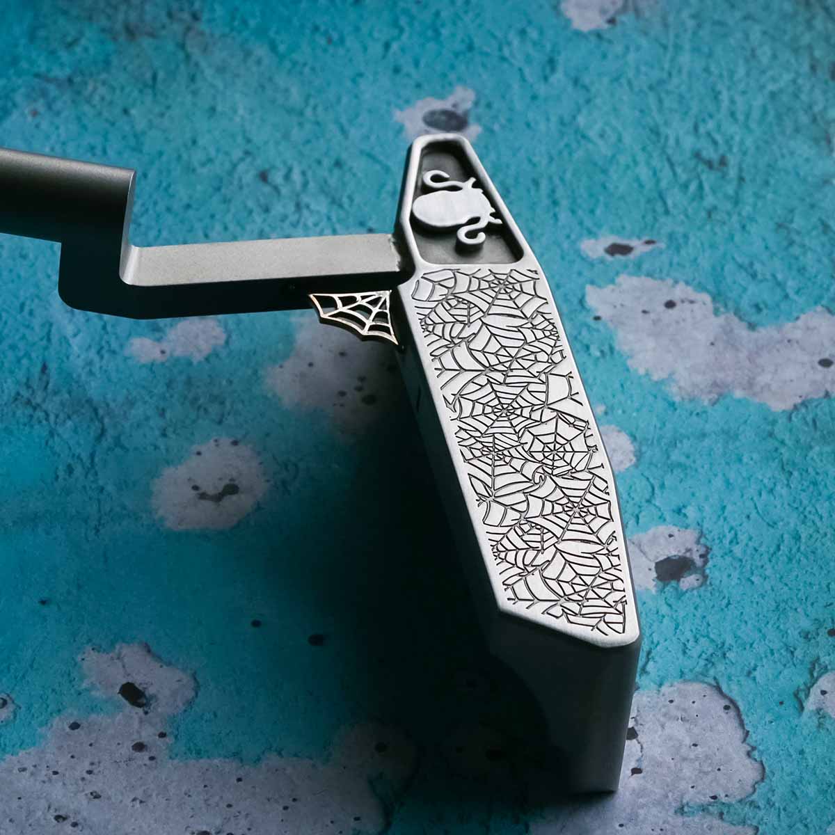 Face view of Arachne stainless steel golf putter with engraved spider pattern and welded Kraken logo inlay near heel