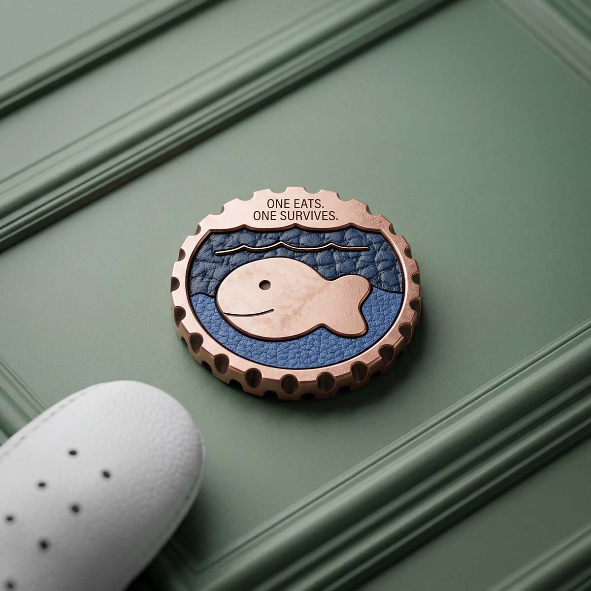 Raised copper goldfish milled proud of a two-tone navy and royal blue leather inlay on a solid copper golf ball marker, with ONE EATS. ONE SURVIVES. milled across the top edge.