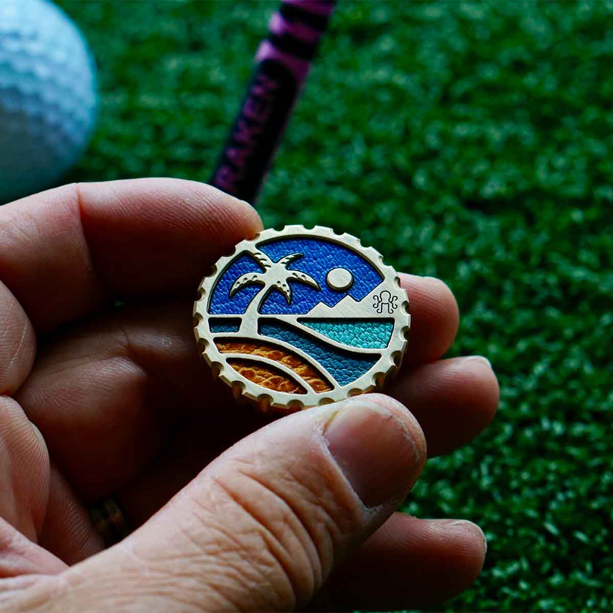 Beachfront Escape brass golf ball marker with raised palm tree, mountain vista, and Italian leather inlays in camel, Tiffany blue, slate blue, and sky blue - Kraken Golf limited edition