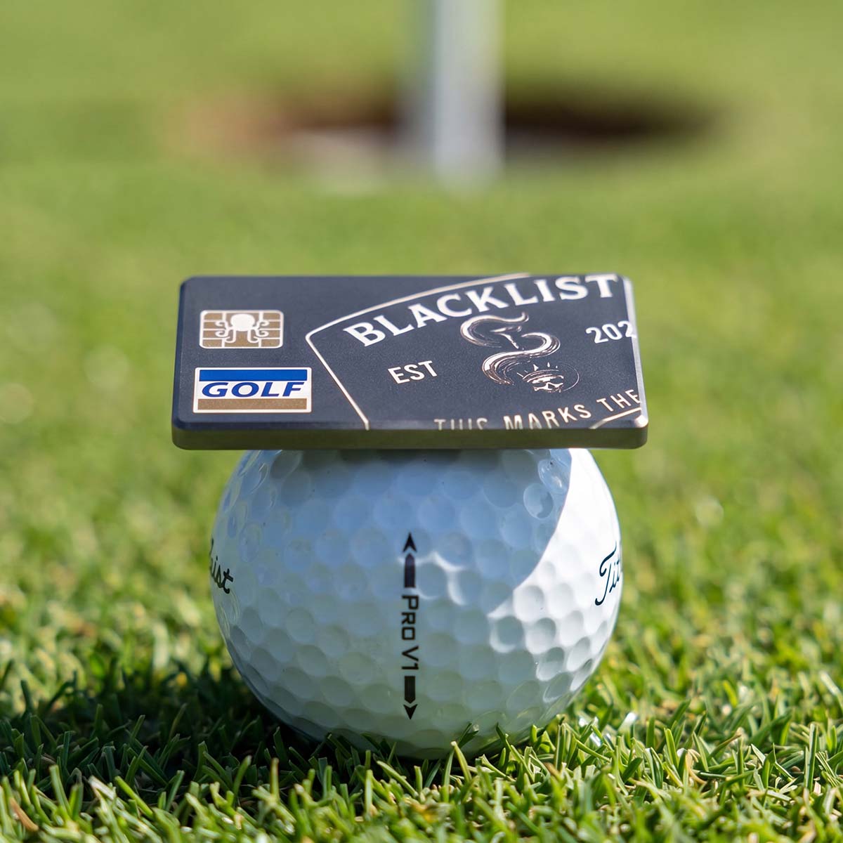 Blacklist Black Card titanium golf ball marker resting on a Titleist Pro V1 ball with the hole and flagstick behind on a putting green
