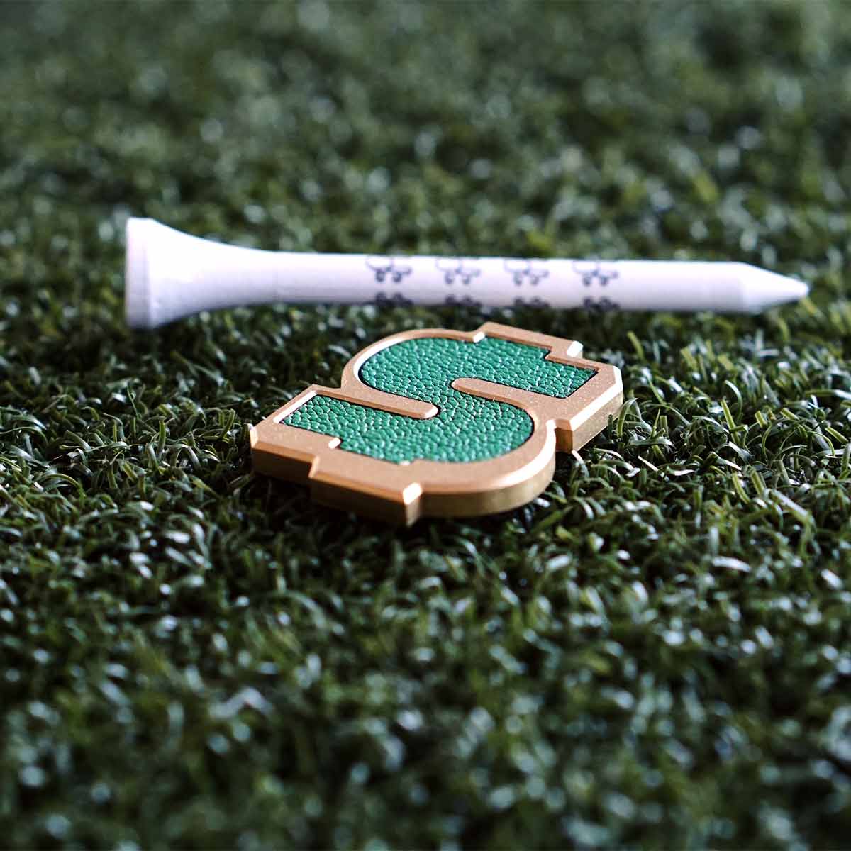 Golf ball marker in the sahpe of a dollar sign on grass with a golf tee in the background