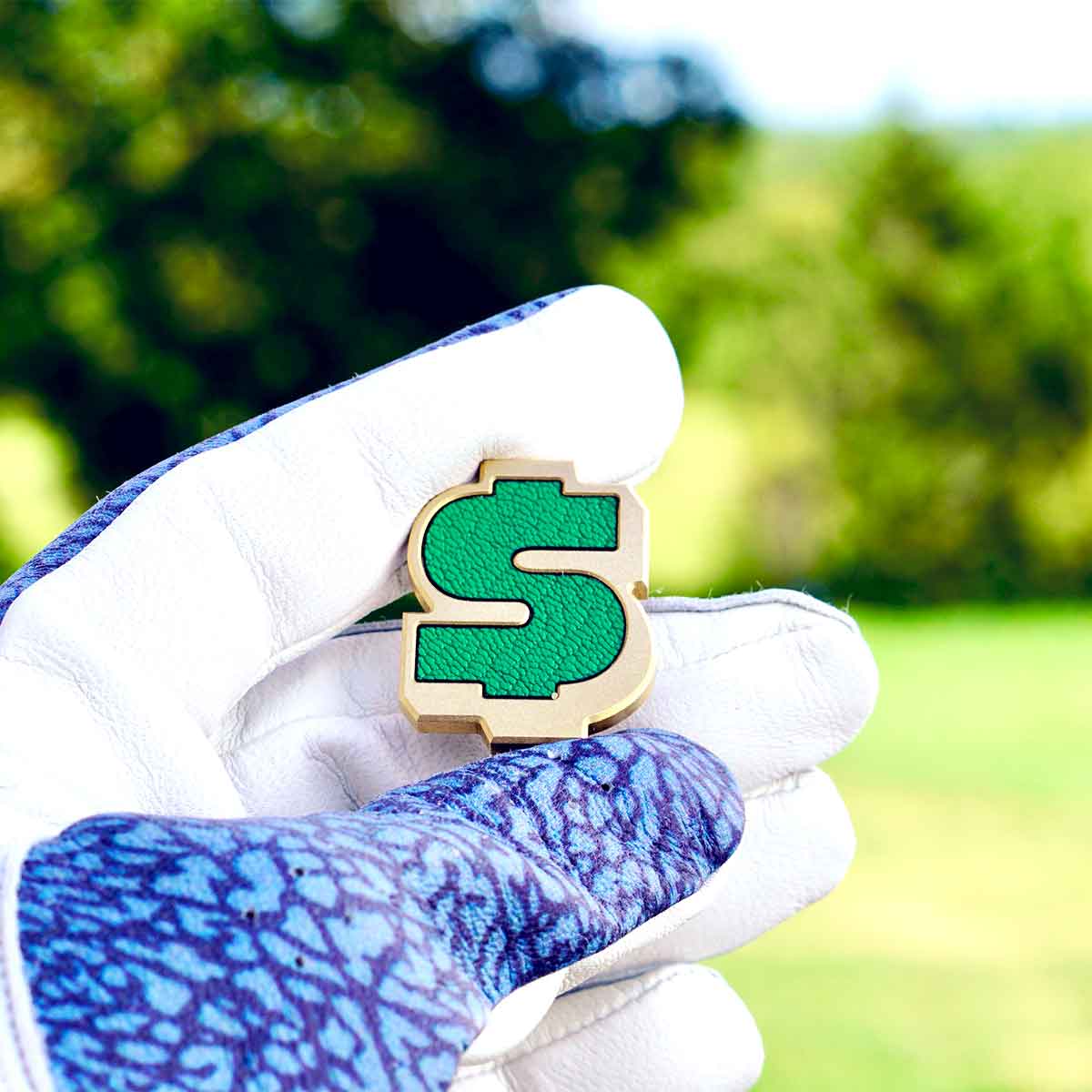 Brass money sign shaped ball marker with green leather inlay heald by a golfer with a golf glove