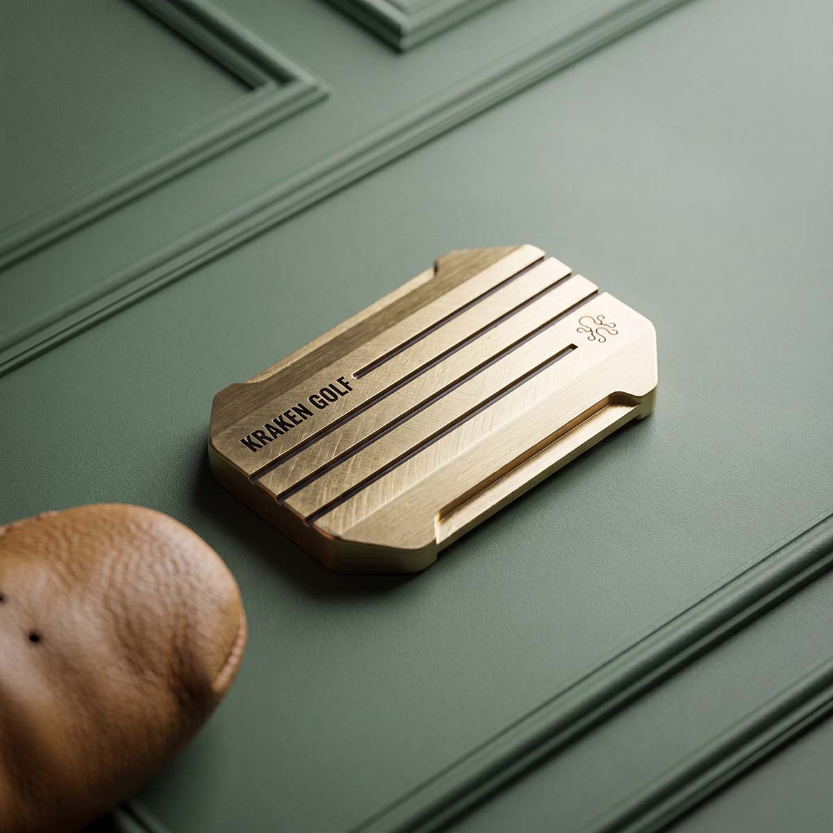 KING COLD brass ball marker reverse — brushed brass with KRAKEN GOLF wordmark and octopus logo, resting on green panel beside a leather headcover