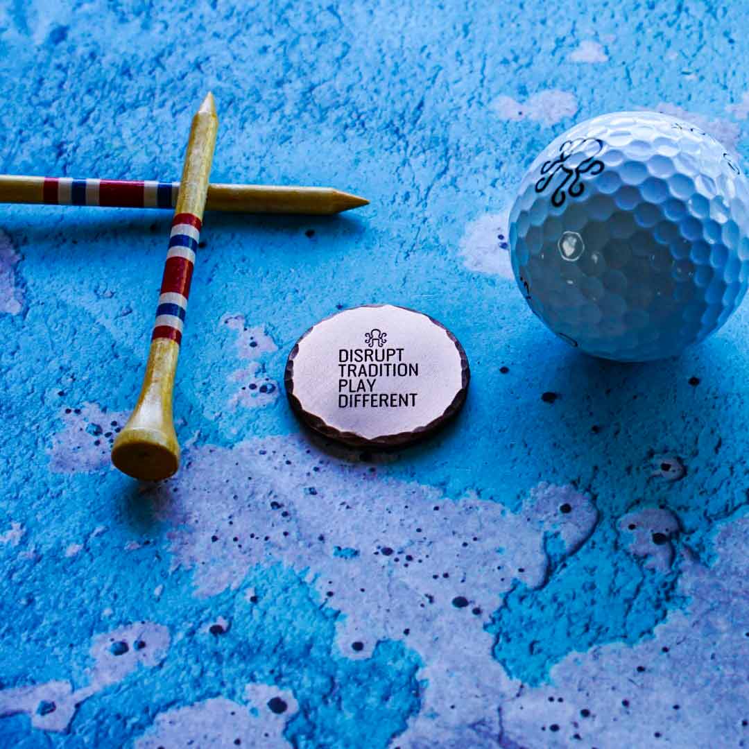 Golf ball, tee, and ball marker with 'Disrupt Tradition Play Different' text on a blue textured surface