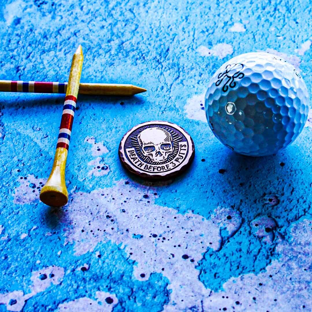 Distressed copper golf ball marker with dark patina finish, raised skull and banner engraving, and hand-hammered edge. Kraken Golf limited edition.