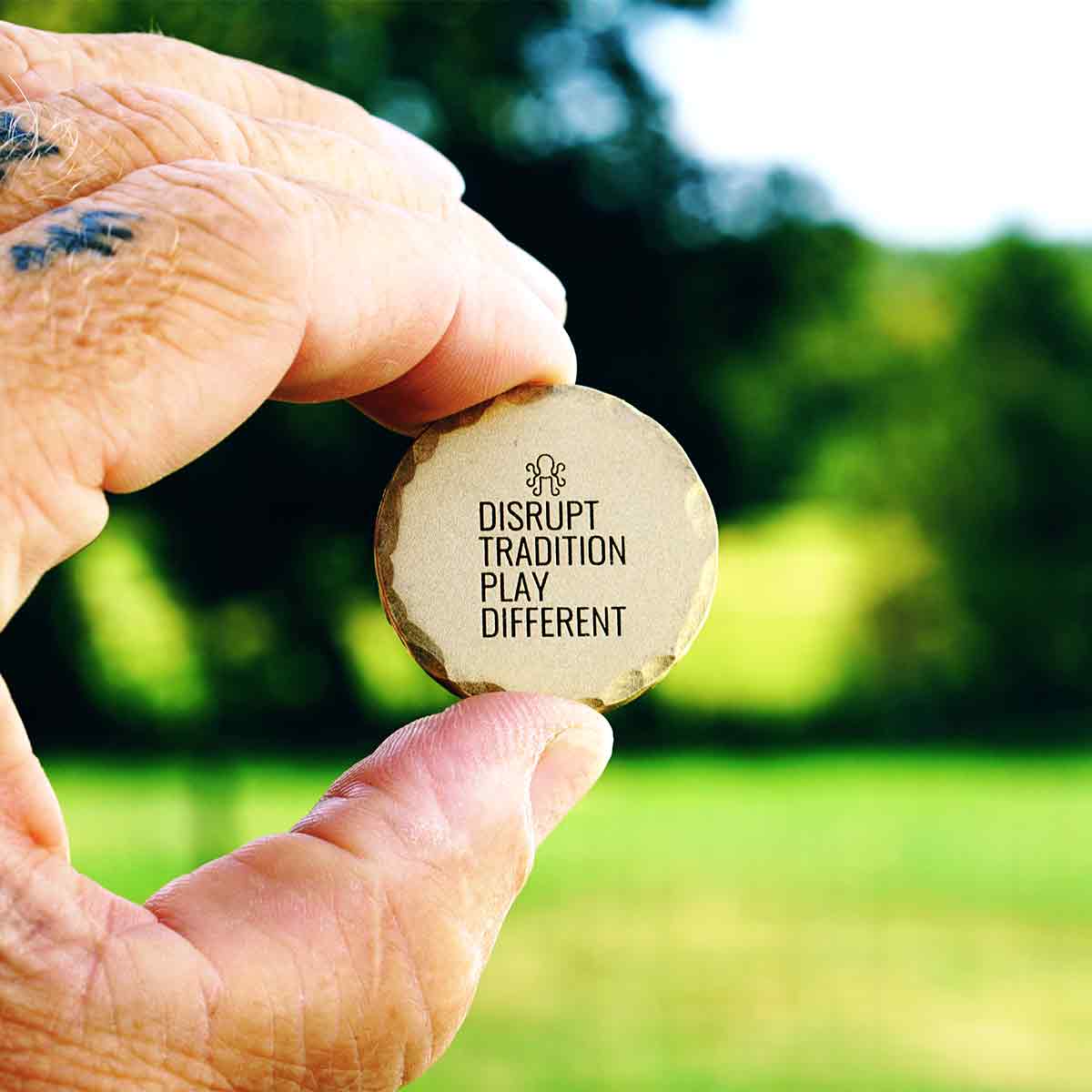 Hand holding a brass ball marker with 'Disrupt Tradition, Play Different' text engraved against a blurred green outdoor background.
