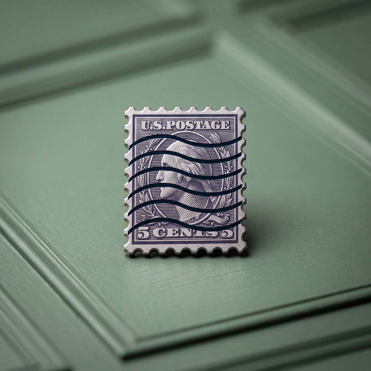 FACE VALUE golf ball marker in blue anodized titanium, shaped like a vintage five-cent postage stamp with a milled perforated edge, profile portrait in an oval, laurels, and hand-cut cancellation lines.
