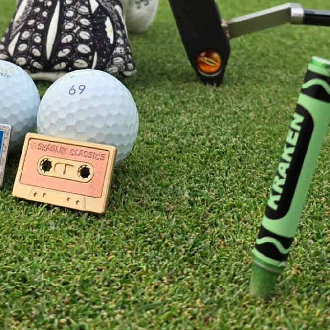 Two golf balls with vintage cassette tape ball marker designs on a golf course, next to a golf club.