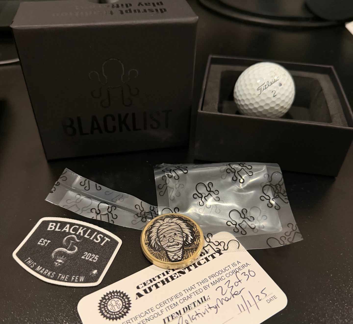 Golf ball, ball marker , and packaging with 'Blacklist' branding on a dark surface.