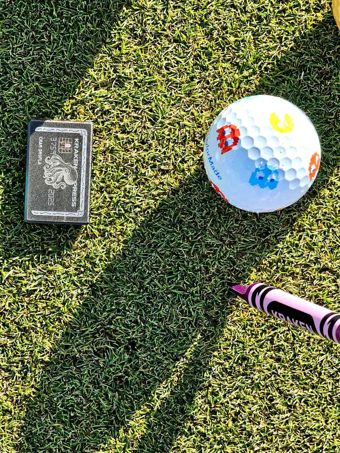 Golf ball with colorful design on grass next to a golf club and a  Kraken credit card ball marker