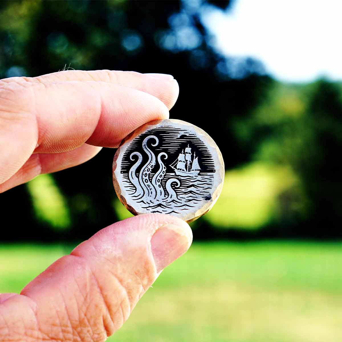 Hand holding a brass golf ball marker with a design of a ship and tentacles in the ocean against a blurred natural background
