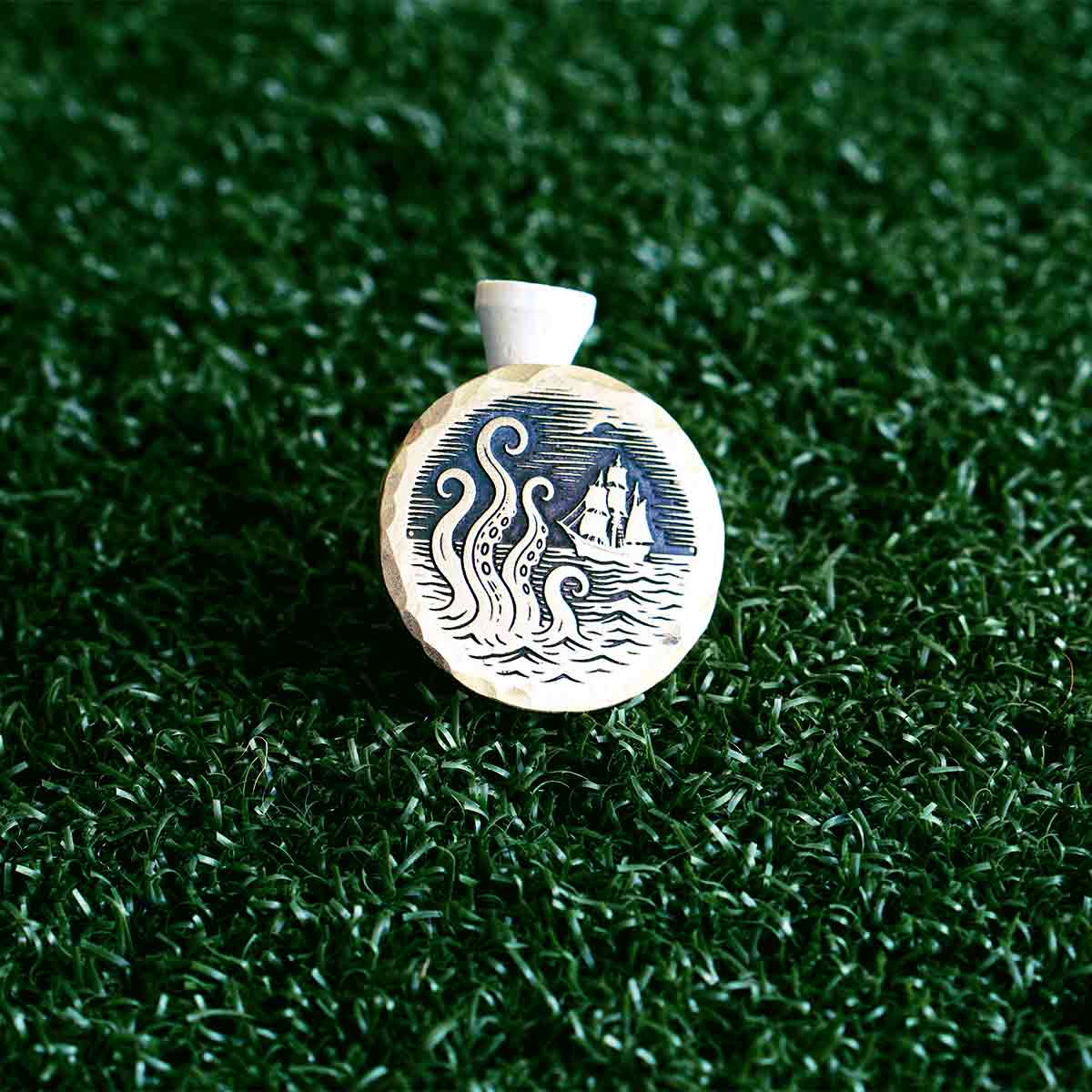 Brass golf ball marker with a ship and octopus design on a green grass background