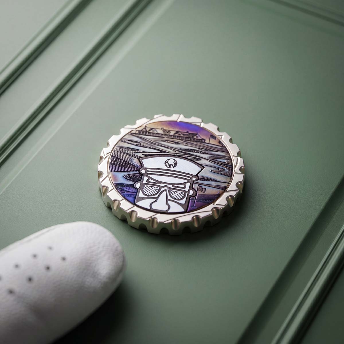 Open Season brass ball marker — stainless steel Captain head and cap inlay over a torched carbon-steel course scene, polished brass face, Southampton 2026