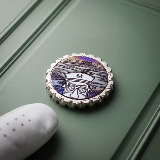 Open Season brass ball marker — stainless steel Captain head and cap inlay over a torched carbon-steel course scene, polished brass face, Southampton 2026