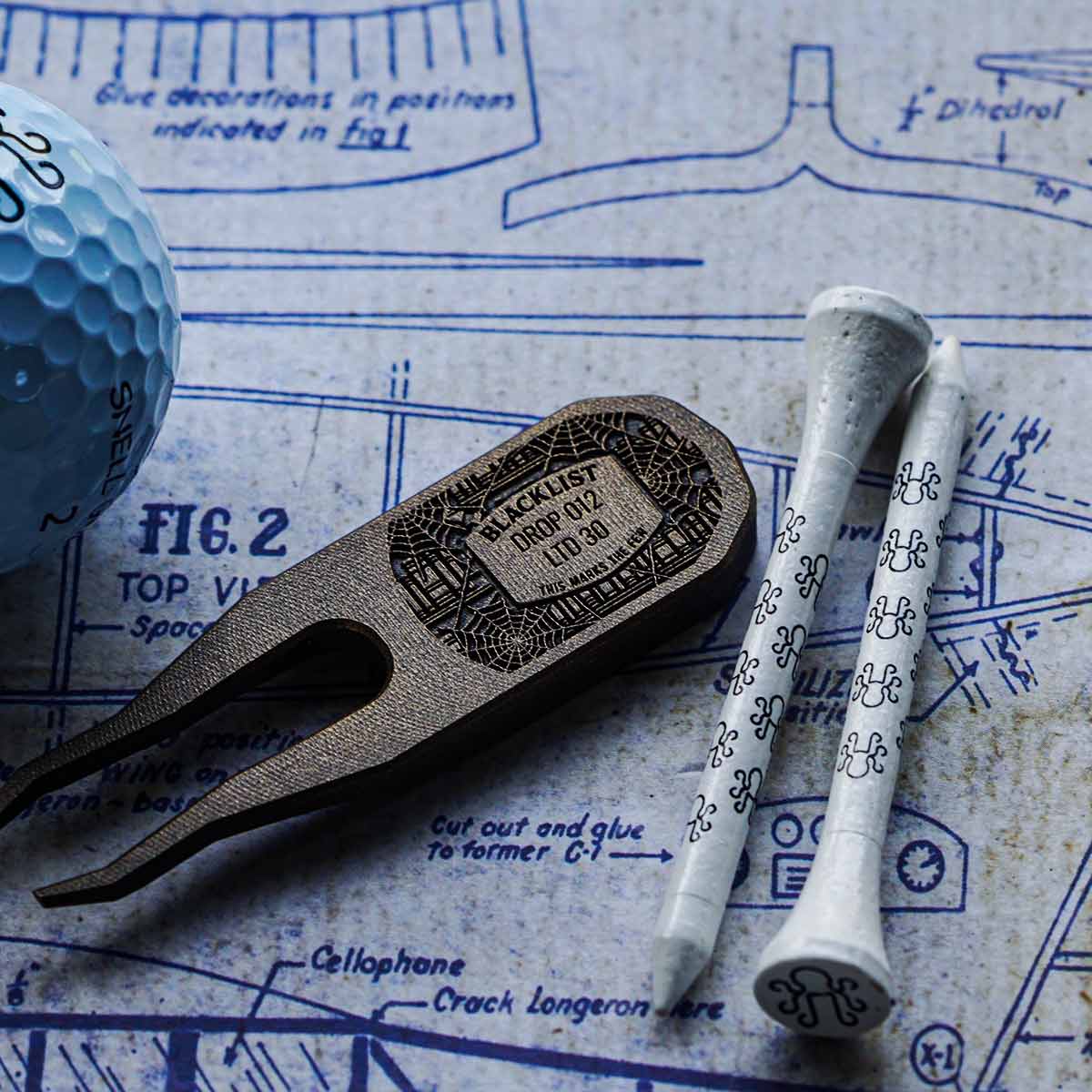 Reverse of brass golf divot tool engraved with Blacklist Drop 013 and This Marks the Few text on cobwebbed bookshelf design