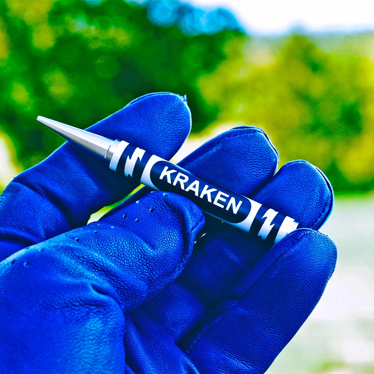 Hand wearing a blue golf glove holding a Kraken crayon pitch tool with a blurred green outdoor background