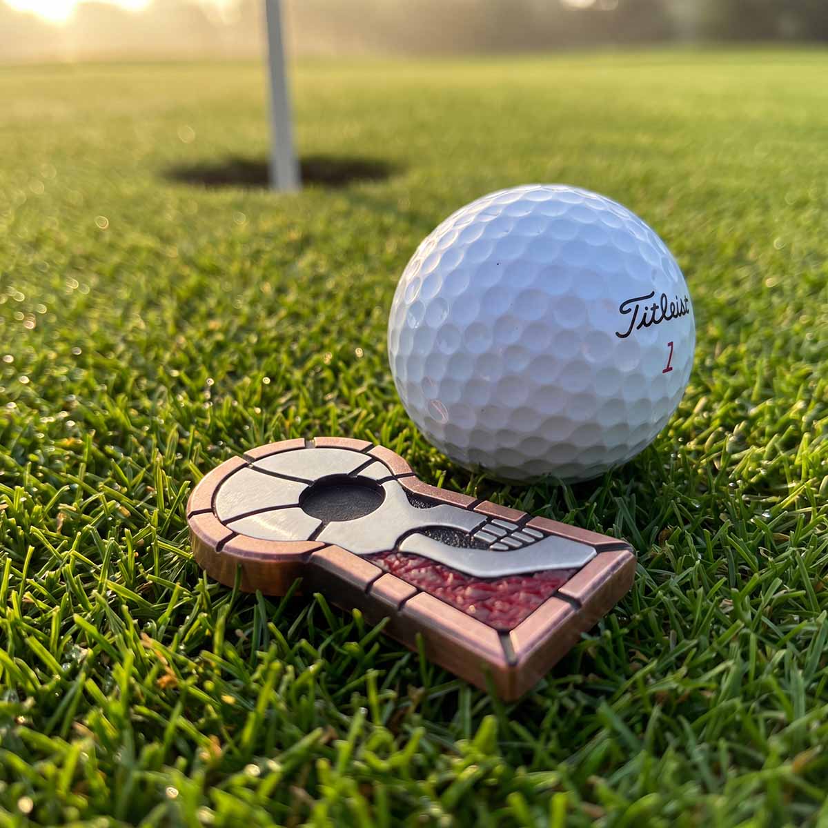 The Keep copper golf ball marker with stainless steel skull inlay beside a golf ball on a putting green at sunrise