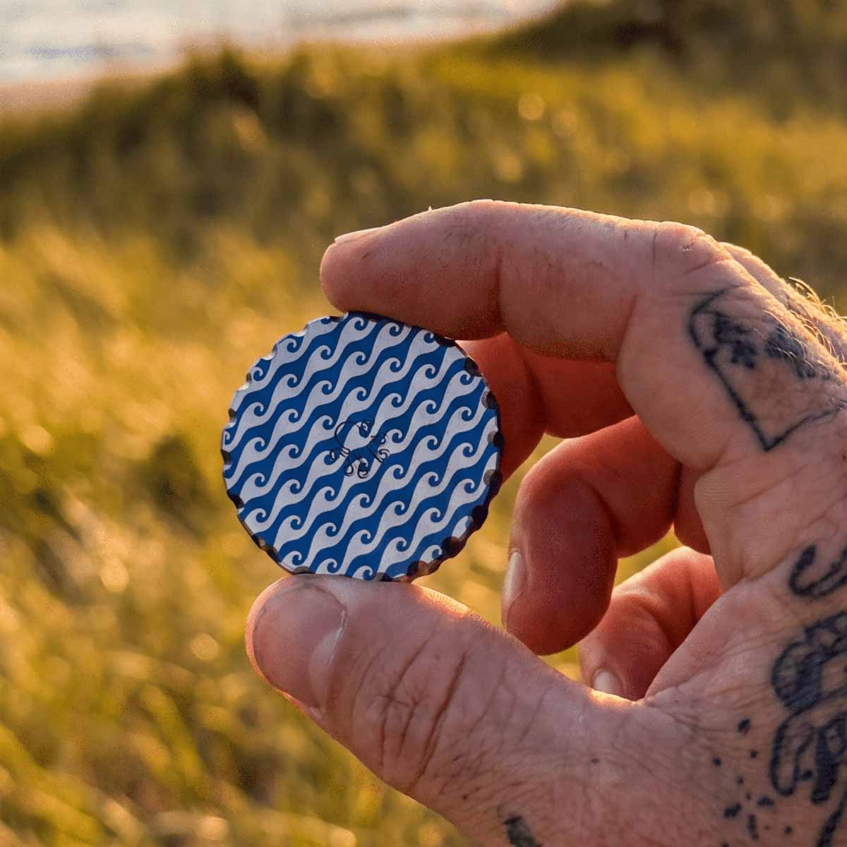 Back of the Kraken Golf Watcher ball marker - centered Kraken logo on a white ocean-wave pattern over anodized blue aluminum, scalloped edge.