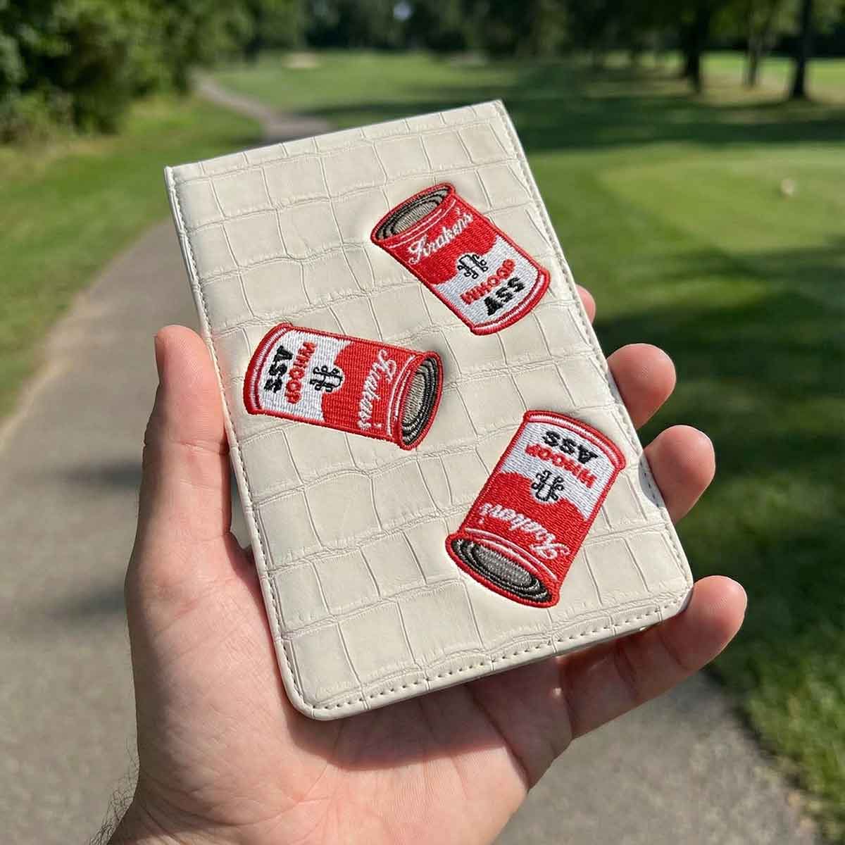 Hand holding a the can of whoop ass golf yardage book with red and white labels against a green outdoor background